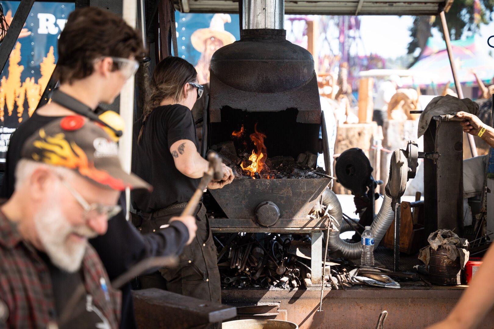 A blacksmith working metal in the forge, orange flames and sparks visible in the forge. Two other blacksmiths watch and smile.