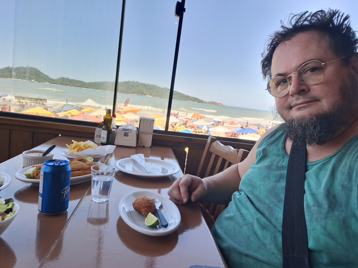 A person sits at a wooden table inside a beachfront restaurant, with the ocean and beach visible through the window behind them. The table holds plates of food, a can of soda, fries and condiments. Outside, the sand is filled with umbrellas, and green hills appear across the water under a clear blue sky.
