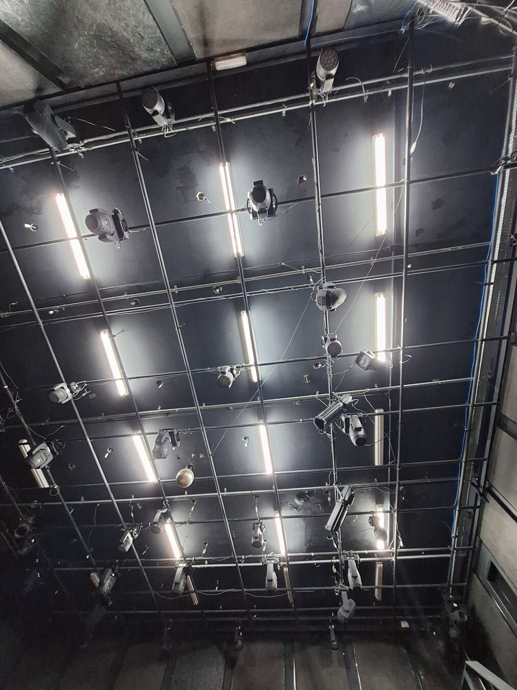 View of a theatre ceiling seen from below, covered with a grid of metal bars supporting multiple stage lights, cables, and fluorescent fixtures. The lights are turned on, creating bright white lines across the dark ceiling, evoking the feeling of a stage waiting for a performance to begin.