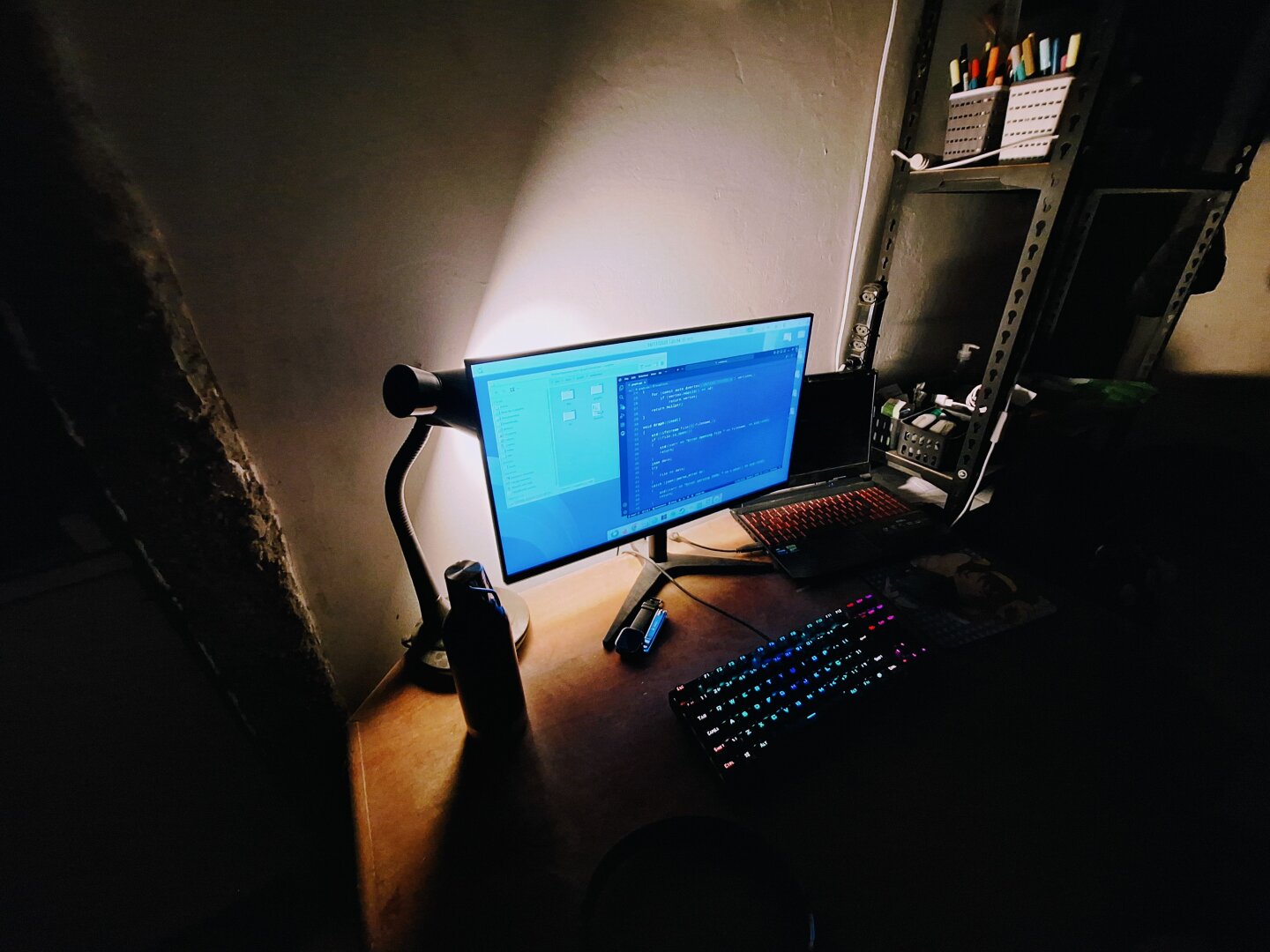 A dimly lit desk with a warm lamp shining onto a wide monitor running a Linux environment styled like the old CDE desktop, giving the scene a retro-tech feeling. The screen shows C++ code, while a rainbow-lit mechanical keyboard glows on the wooden surface. A laptop with an orange backlit keyboard sits beside it, and shelves with pens and tools rise to the right. A water bottle rests near the lamp, and the room around the desk fades into shadow.