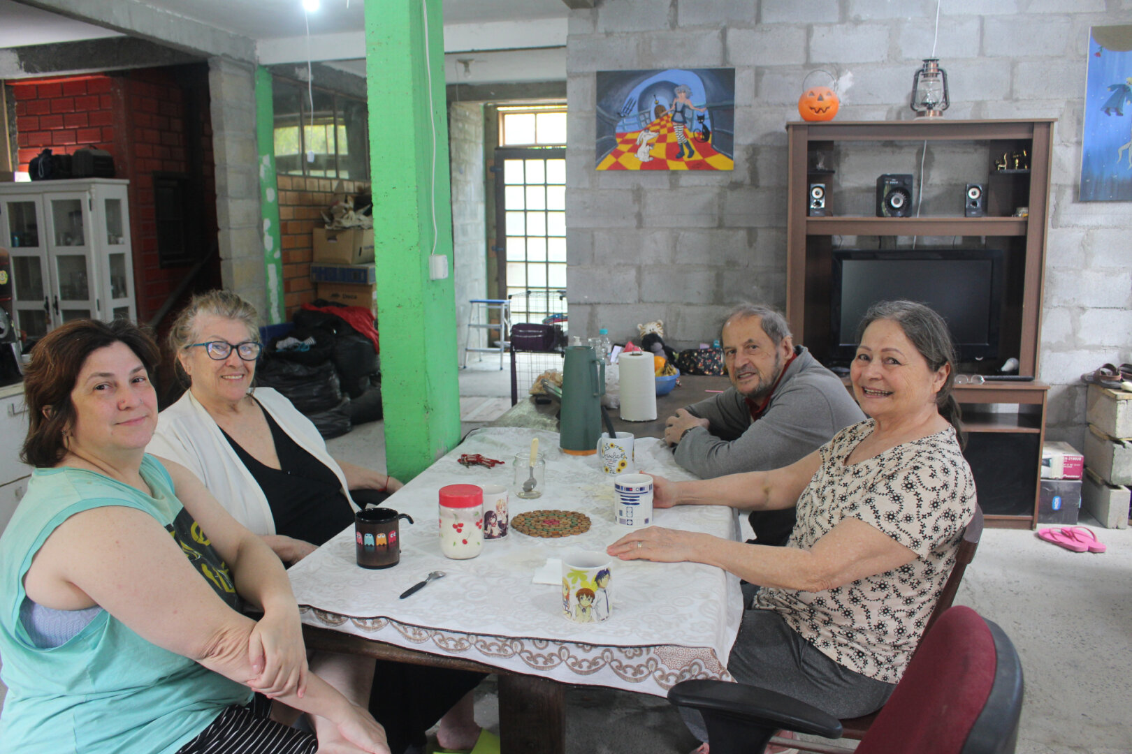 Four people sit around a table in a cozy, rustic room with unfinished brick and concrete walls. They smile warmly toward the camera, mugs and a thermos on the table suggesting a shared coffee moment. The space feels homely and lived-in, decorated with colorful paintings and small personal touches.