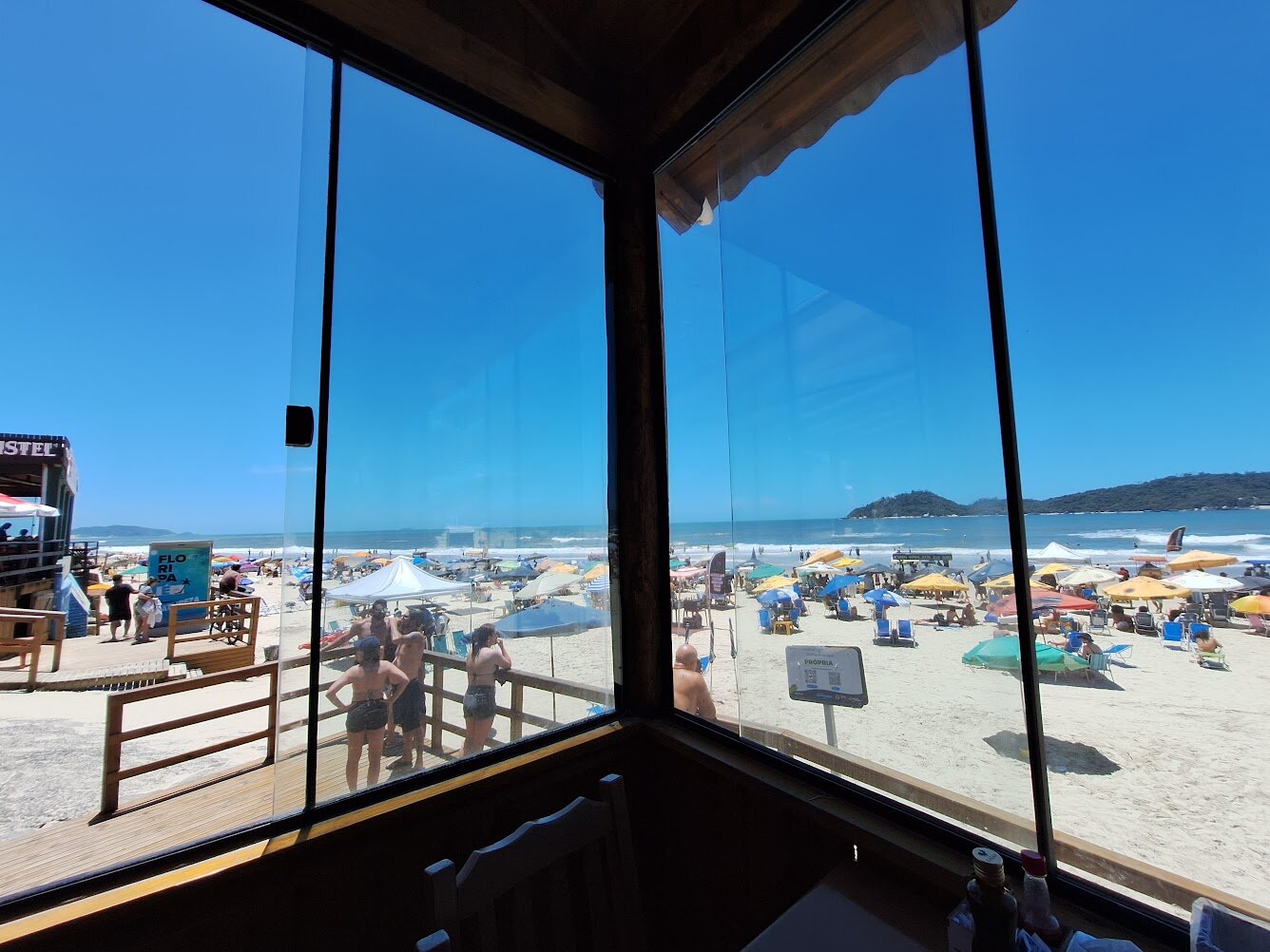 A photo taken from inside a beachfront restaurant, looking out through large glass windows. Outside, the beach is crowded with colorful umbrellas, people walking on the sand and standing near wooden walkways. The sky is bright blue and the ocean waves roll in calmly. Distant green hills rise at the edge of the horizon.