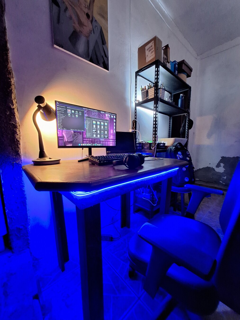 A small workspace glowing with deep blue light, where an old wooden desk holds an wide monitor running a purple-toned Linux system. A laptop, a mechanical keyboard and headphones rest like quiet tools of a maker. A warm lamp brushes the wall with a soft golden halo. On a metal shelf in the background, boxes, pens and small objects wait in careful disorder. The scene feels half workshop, half digital sanctuary, a place where code, art and imagination meet in the dim afterlight of a future that never arrived but still insists on being dreamed.