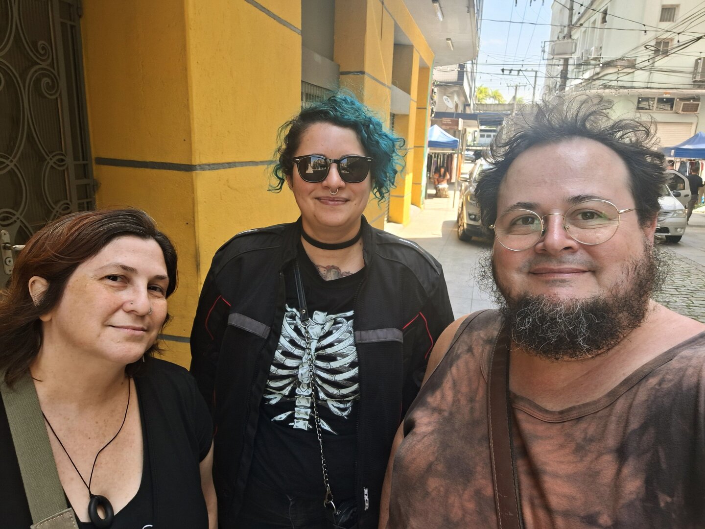 Three friends standing close together on a bright street, smiling at the camera. Behind them are yellow buildings, parked cars and small blue-tented market stalls.