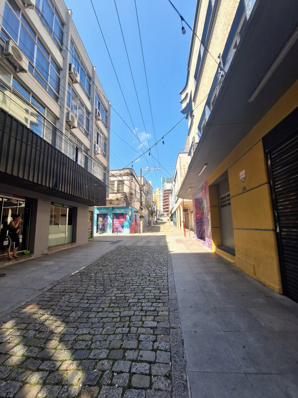 A mostly empty city street on a sunny afternoon. A cobblestone road runs uphill between buildings on both sides. Some walls feature colorful murals. All storefronts are closed, and strings of lights hang across the street.