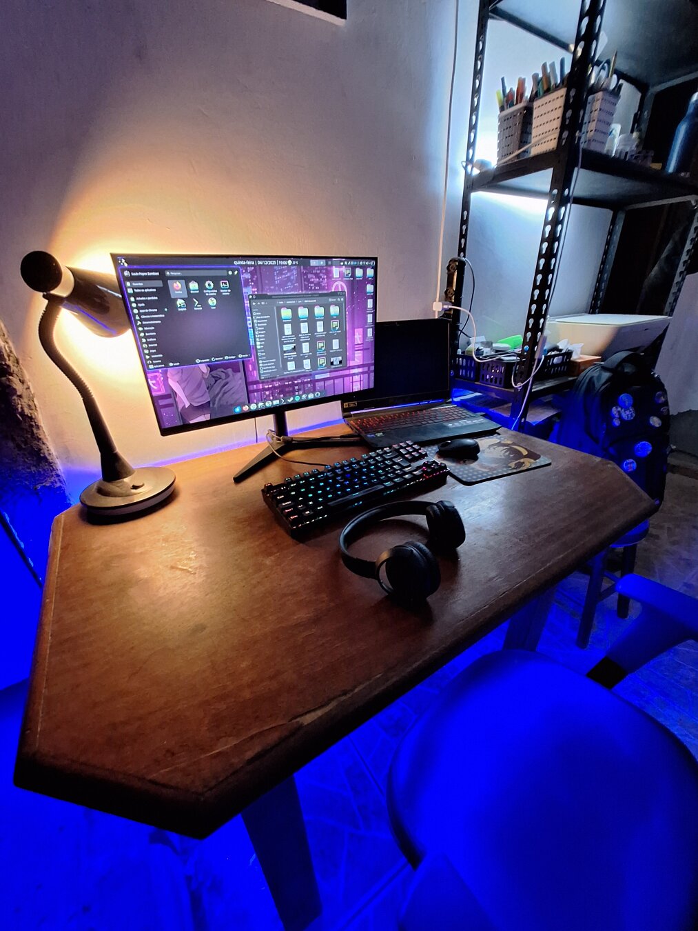 A small workspace glowing with deep blue light, where an old wooden desk holds an wide monitor running a purple-toned Linux system. A laptop, a mechanical keyboard and headphones rest like quiet tools of a maker. A warm lamp brushes the wall with a soft golden halo. On a metal shelf in the background, boxes, pens and small objects wait in careful disorder. The scene feels half workshop, half digital sanctuary, a place where code, art and imagination meet in the dim afterlight of a future that never arrived but still insists on being dreamed.