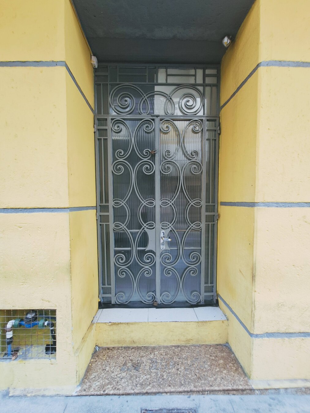 A yellow building entrance with a metal gate decorated with swirling, ornate patterns. Behind the gate, frosted glass panels soften the view of the interior. The doorway sits slightly elevated, with a worn doormat at the step. To the left, a small grated opening reveals exposed water pipes. The scene feels both ordinary and quietly expressive, as if the curves of the gate try to soften the strict geometry of the yellow walls.