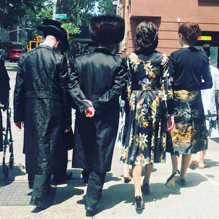 Four people, back to back, wearing traditional Orthodox Jewish clothing, walking through the Williamsburg neighborhood in Brooklyn, New York.