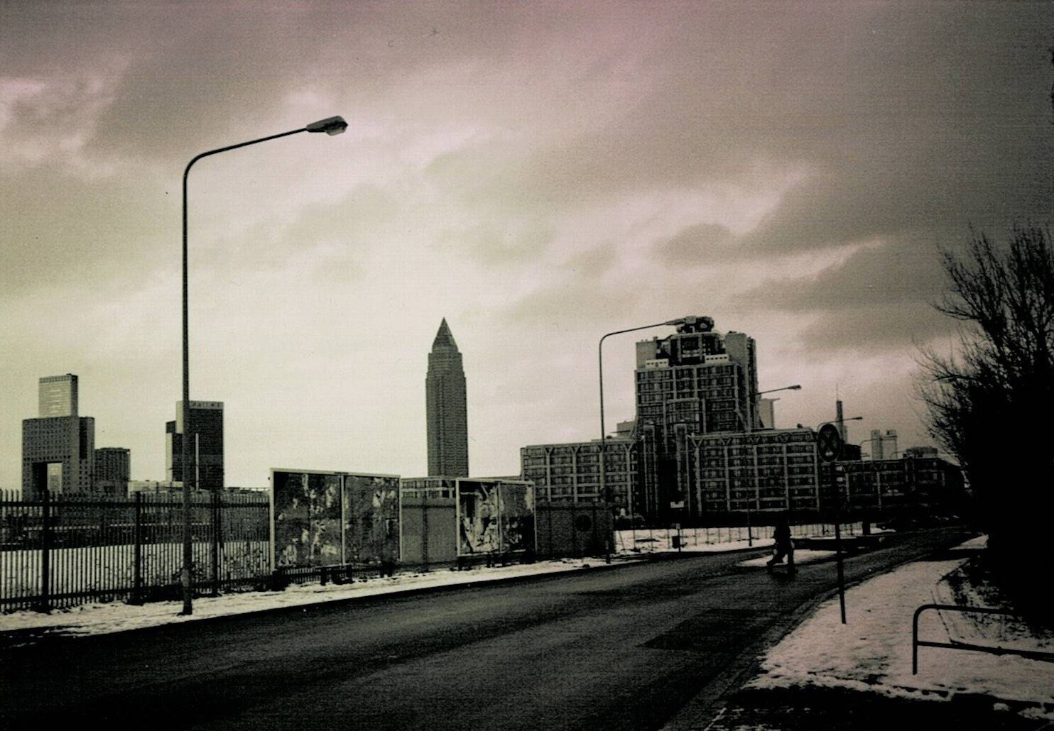 a street, maybe in Frankfurt/Main, a little snow is left. 
There is a fence, there is a streetlight, there is a person in a distance, crossing the street, no cars. 
Further in the distance, you can see some big towers as a kind of fractured skyline. 
It is a cloudy day.