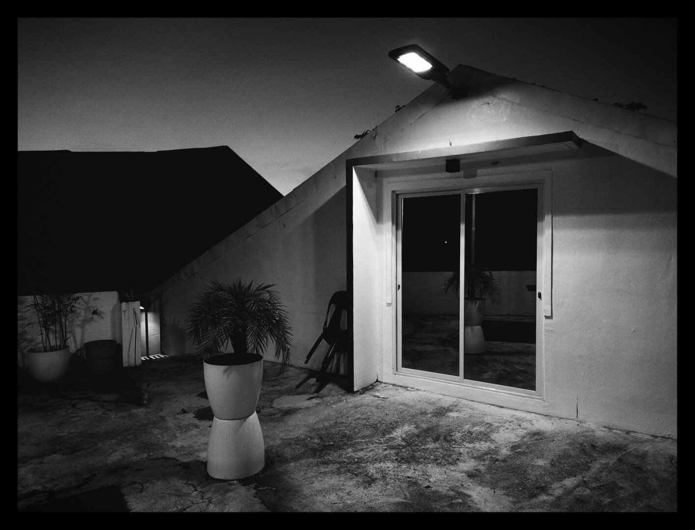 A black and white photo shot on a house terrace, showing a potted tree and a sliding glass door in the foreground, with a chair next to it near the wall. The terrace is lit up by a solar rectangular light above the glass door.

The back of the structure of the warehouse is in the background. This picture is taken during evening.