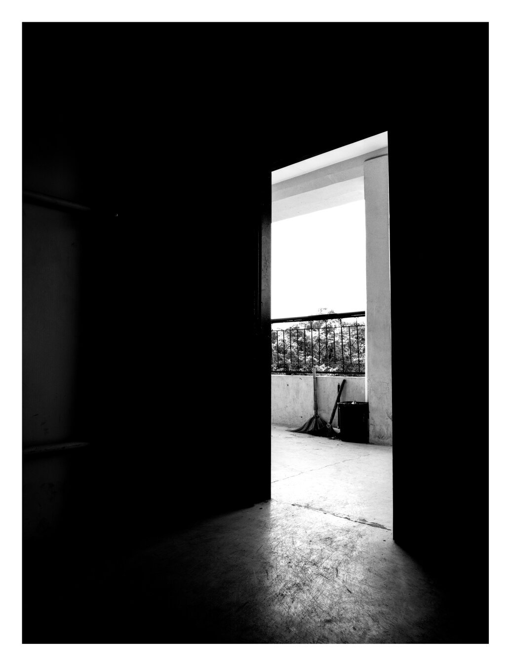 A black and white photograph depicting an open doorway leading to a corridor with an outside view with trees' leaves visible. Two brooms and a trash can are also visible, on the corridor.