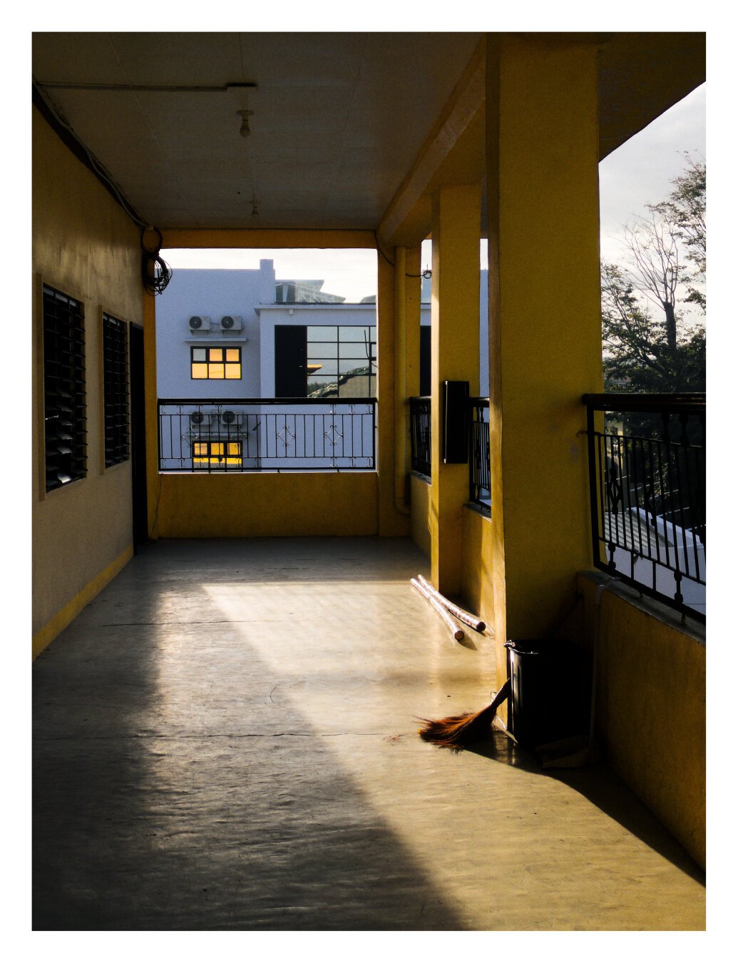 An outdoor shot of the end of the third floor corridor of the school building, with the sunlight casting on the floor.

The trash can is in the foreground, visible on the right side of the walkway, as well as the broom leaning against the wall near the trash can.