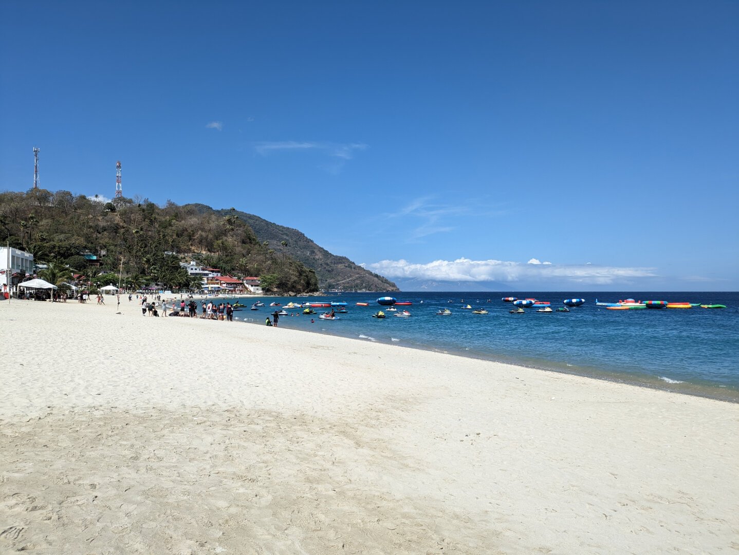 The photo captures a picturesque scene of White Beach in Puerto Galera, Oriental Mindoro, Philippines. The beach stretches across the horizon, adorned with pristine white sand and lapped by turquoise waters. Colorful boats bob gently in the distance, adding a vibrant touch to the scene. A lush green hillside rises behind the beach, providing a backdrop of natural beauty. The sun shines brightly, casting a warm glow over the entire landscape. - Google Gemini