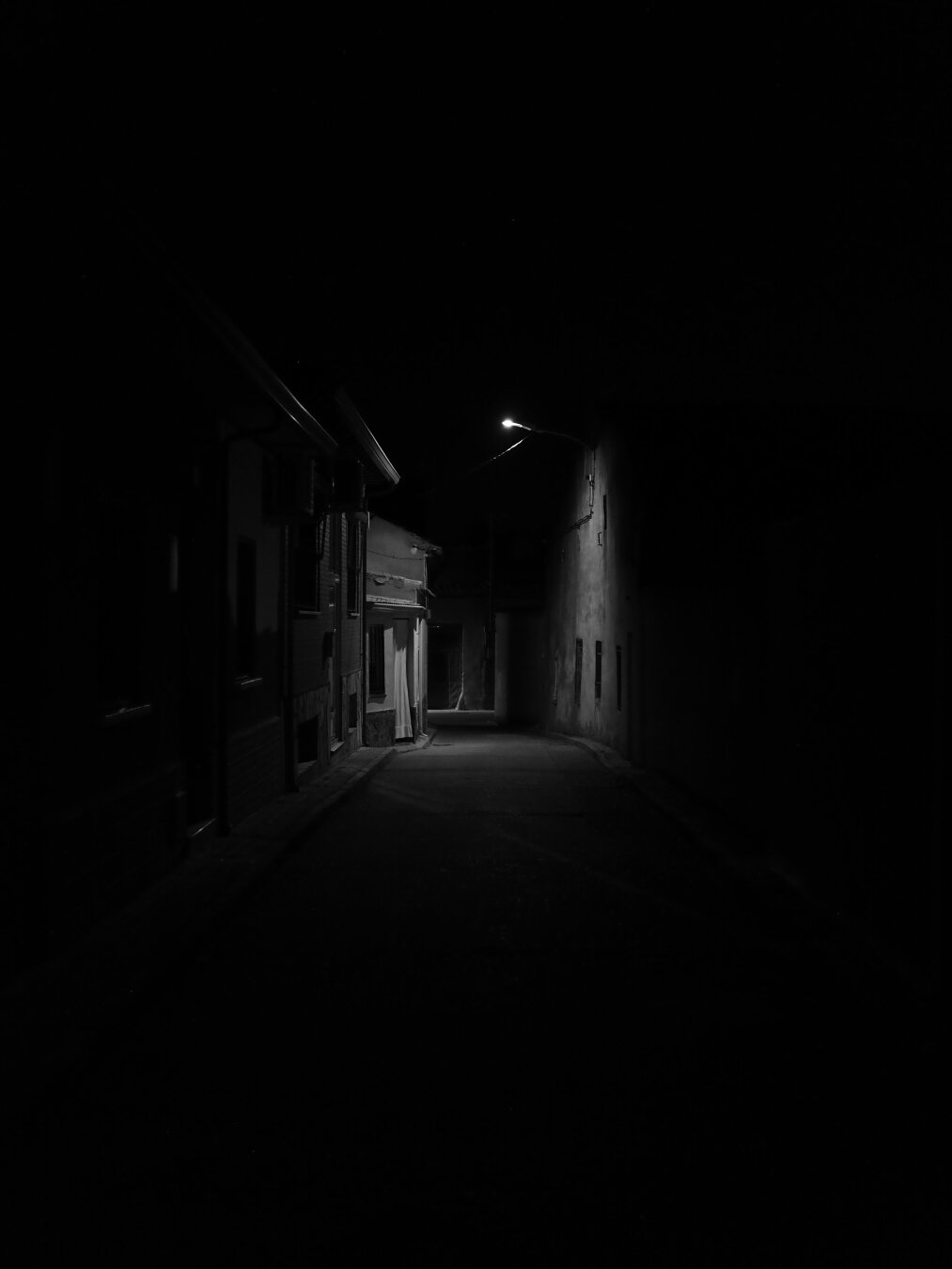 Calle estrecha de noche, en blanco y negro, prácticamente sumida en la oscuridad. Una farola ilumina parcialmente las fachadas y el tramo central de la calle, creando fuertes contrastes entre luces y sombras.