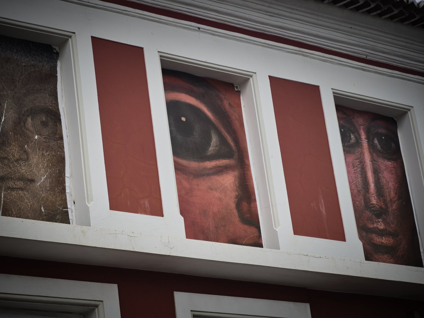 Fachada de un edificio en Angra do Heroísmo, Azores, fotografiada en contrapicado. Entre molduras blancas y paños rojizos aparecen grandes imágenes de un ojo y parte de un rostro, integradas en los huecos superiores del edificio.