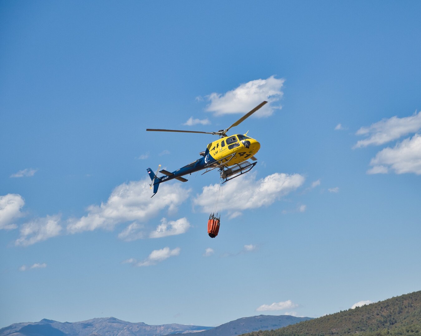 Yellow and blue firefighting helicopter flying away with an orange water bucket suspended beneath it, surrounded by a wide blue sky with scattered clouds and distant mountains below.