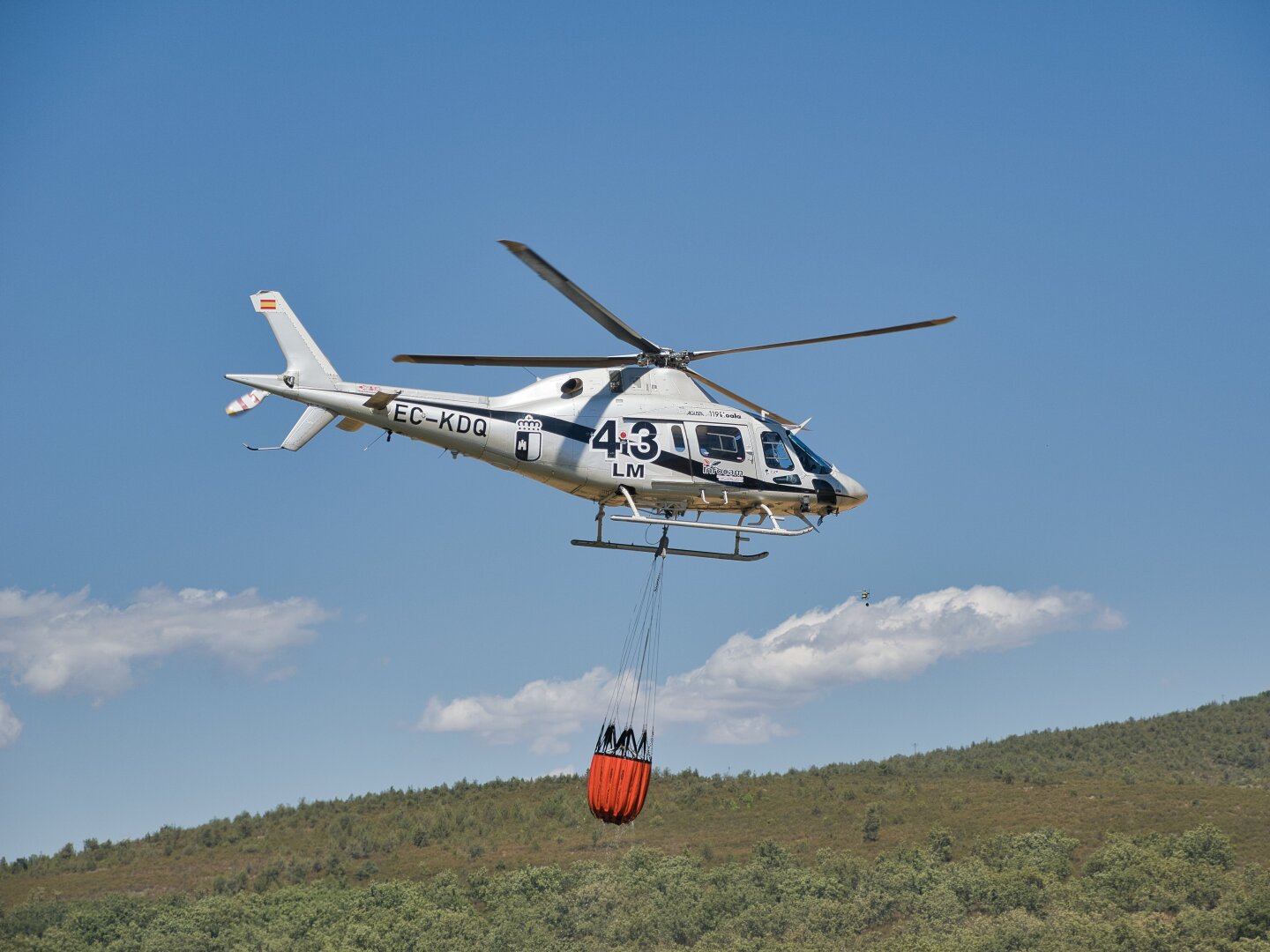Close side view of a white firefighting helicopter flying with an orange water bucket suspended beneath it. The aircraft stands out against a blue sky, with low wooded hills below.