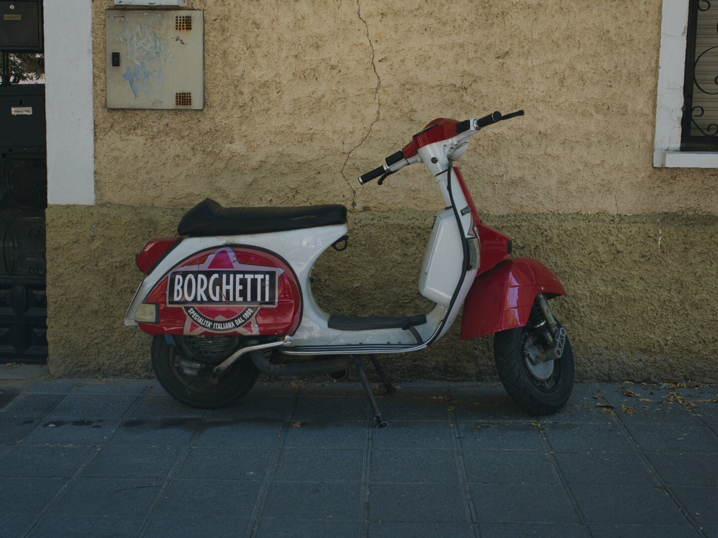 A red-and-white Vespa scooter parked beside a pale wall on a quiet street. The scooter is shown almost in profile, with its rounded bodywork, mirrors and rear storage box clearly visible. Warm pavement and the simple wall create a clean, uncluttered background that makes the red details stand out.
