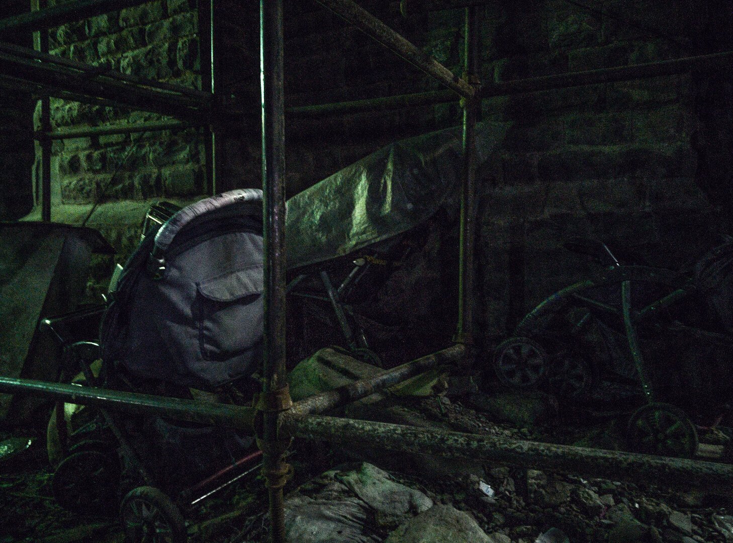 two baby prams entangled between metal construction scaffolds. the scene is bathed in green light at night time and the image features digital noise.