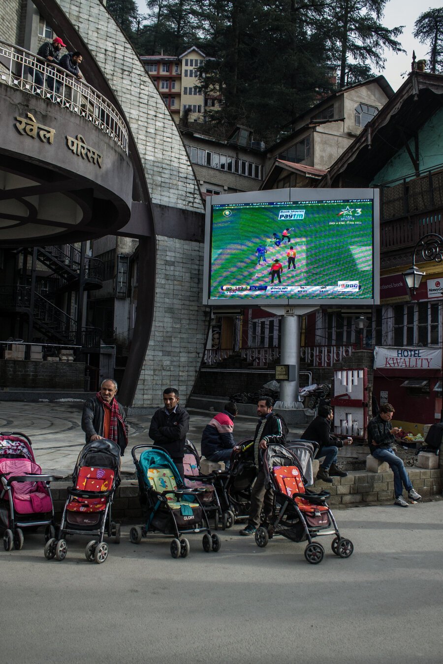 A big screen mounted into a thick pole displays a cricket game in the background. To the left of this screen is an arch of a building, half visible in the image. There is a balcony near this arch where two people are standing and watching the big screen. In the foreground, there are various people standing and sitting near baby strollers, looking away from the big screen.