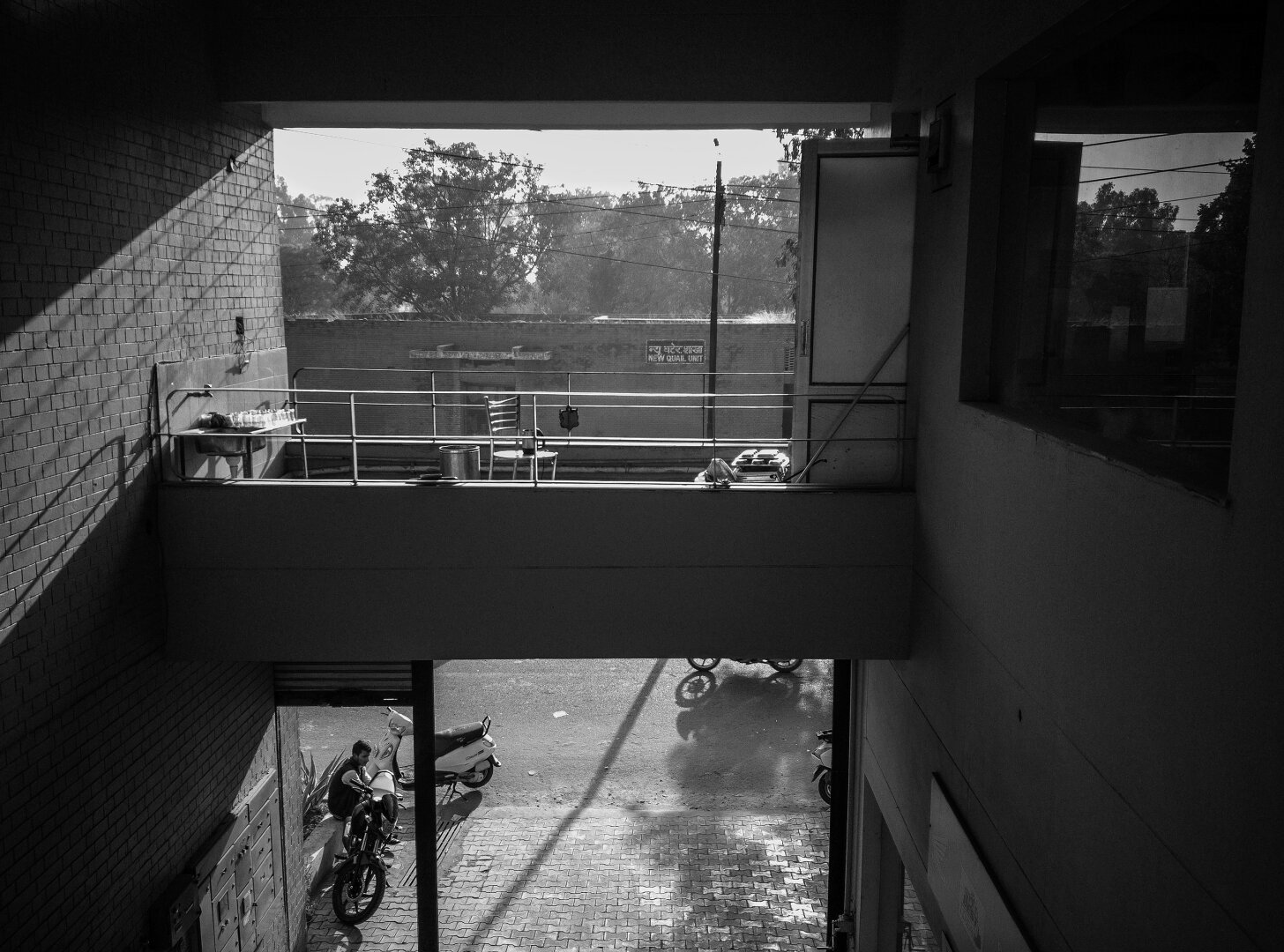 A monochrome image of a balcony in the middle, with a sink and washed glasses to the left of the balcony, a seat in the middle of it and a door to the right of it. The sunlight streaming from the front of the balcony casts a slanted shadow on the left wall of the balcony. Beneath the balcony are some motorbikes and a scooter with someone sitting on a ledge on the roadside.