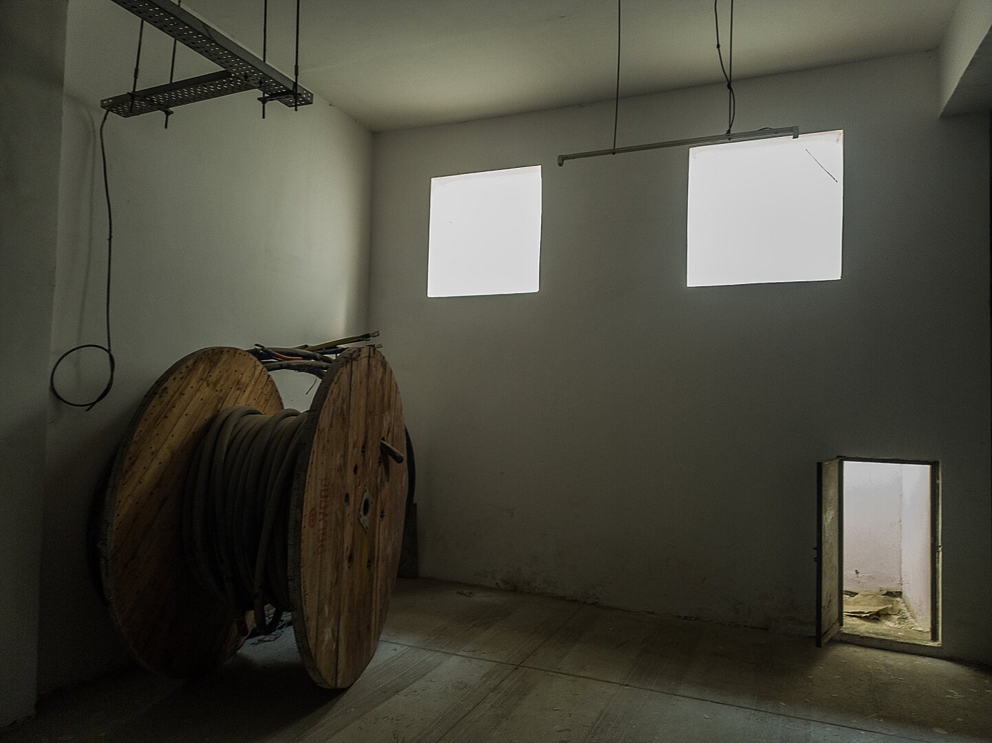 A spool of thick underground wire wrapped around a large wooden spool at the left of the image. In front of this spool is a wall with two unshuttered, open windows blaring light into the room. To the image bottom right on the same wall is a shuttered passage with an open metal door also letting some light in.