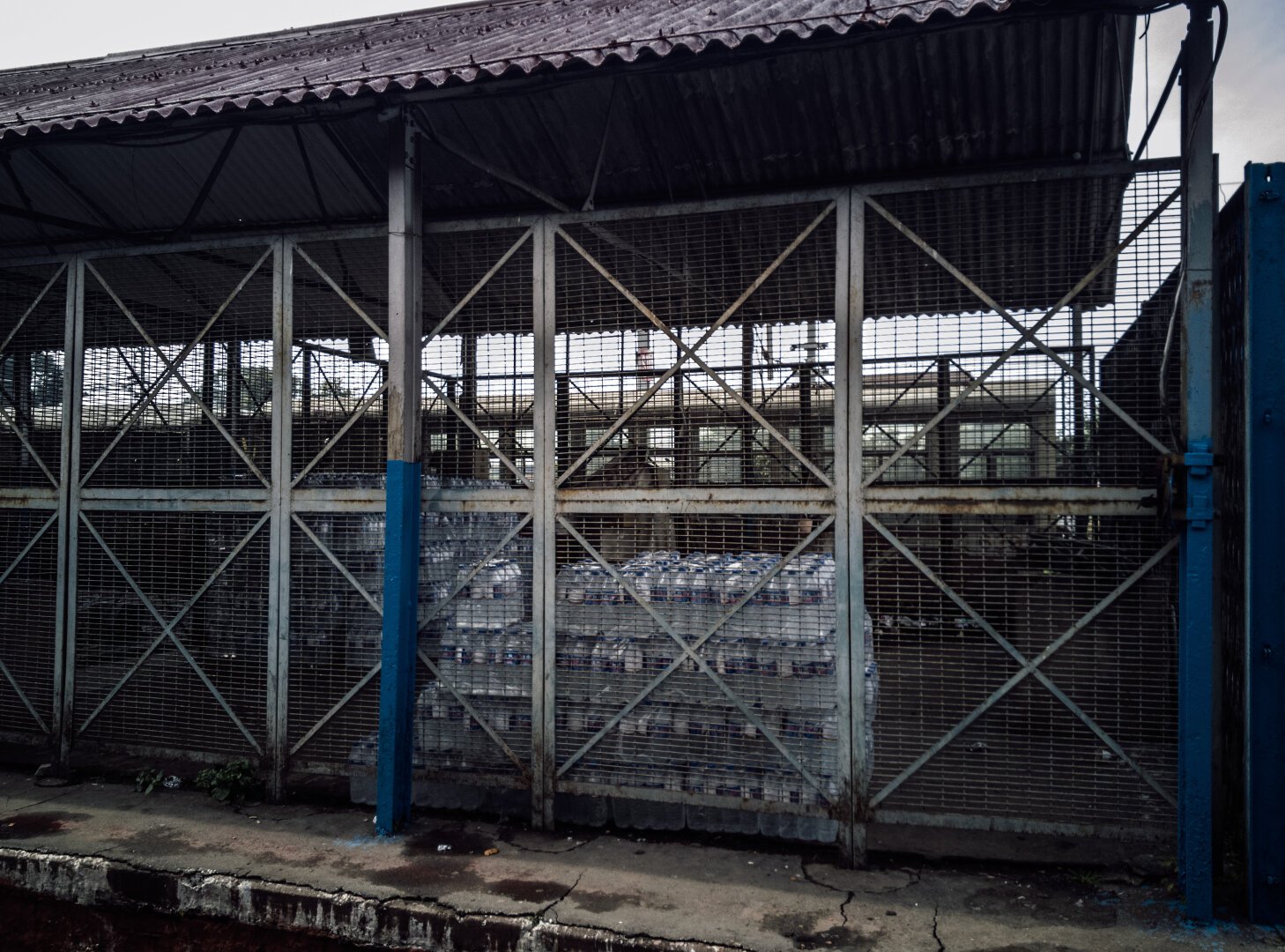 A photo of a locked metal shed containing multiple pallets worth of plastic water bottles packed in plastic packaging together. in the background, behind the locked shed, a train carriage is visible.