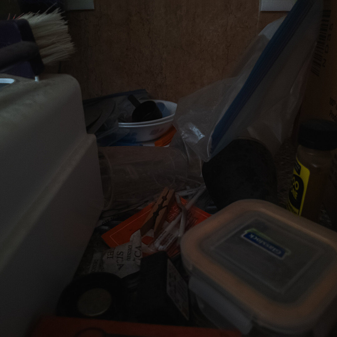 A random assortment of items strewn about a surface. The image is taken in a low light condition with some items barely visible.

To the left, there is a white box with a ridge and a white bristled plastic brush on top of it. In the middle, at the back, there is a stack of white bowls containing a scoop and straddling a flat ziplock bag. In the middle, towards the front, there are the following items: a wooden clothes clip, plastic straws wrapped in plastic bags, a silica gel packet, a circular bottle cap, a square glass container with a clip on lid that has an orange gasket visible. To the right, there is another ziplock bag and a bottle of glue remover.
