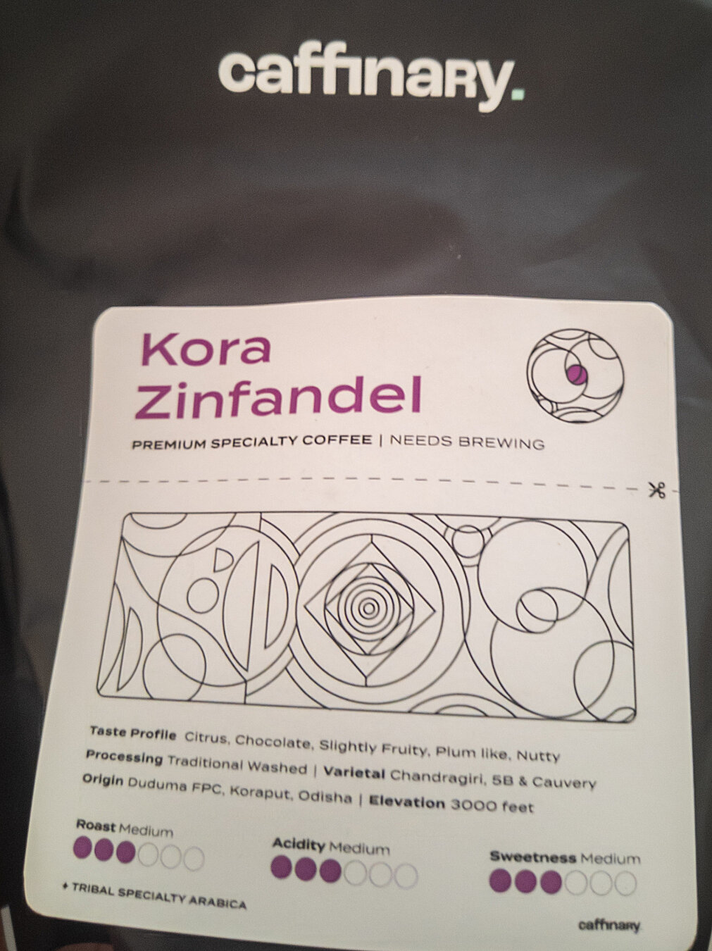 A black coffee bag with a white "Caffinary" logo on top with a teal square period at the end.

Below it, there's a white rectangular label with the following details:
"Kora 
Zinfandel (in maroon text) (a circular graphic with circular geometric outlines within it)

PREMIUM SPECIALTY COFFEE | NEEDS BREWING

(A rectangular graphic with various circular and rectangular geometric outlines within it)

Taste Profile Citrus, Chocolate, Slightly Fruity, Plum Like, Nutty

Processing Traditional Washed | Varietal Chandragiri, 5B & Cauvery

Origin Duduma FPC, Koraput, Odisha | Elevation 3000 feet


Roast Medium    Acidity Medium     Sweetness Medium

TRIBAL SPECIALTY ARABICA      (Caffinary logo)
"