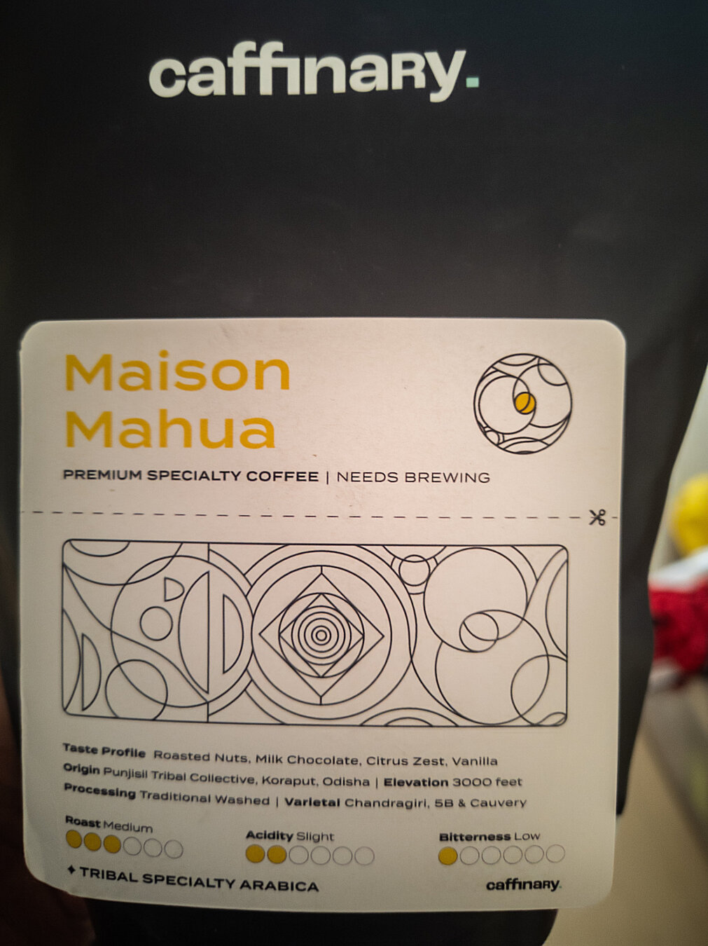 A black coffee bag with a white "Caffinary" logo on top with a teal square period at the end.

Below it, there's a white rectangular label with the following details:
"Maison 
Mahua (in yellow text) (a circular graphic with circular geometric outlines within it)

PREMIUM SPECIALTY COFFEE | NEEDS BREWING

(A rectangular graphic with various circular and rectangular geometric outlines within it)

Taste Profile Roasted Nuts, Milk Chocolate, Citrus Zest, Vanilla
Origin Punjisil Tribal Collective, Koraput, Odisha | Elevation 3000 feet
Processing Traditional Washed | Varietal Chandragiri, 5B & Cauvery

Roast Medium    Acidity Slight     Bitterness Low

TRIBAL SPECIALTY ARABICA      (Caffinary logo)
"