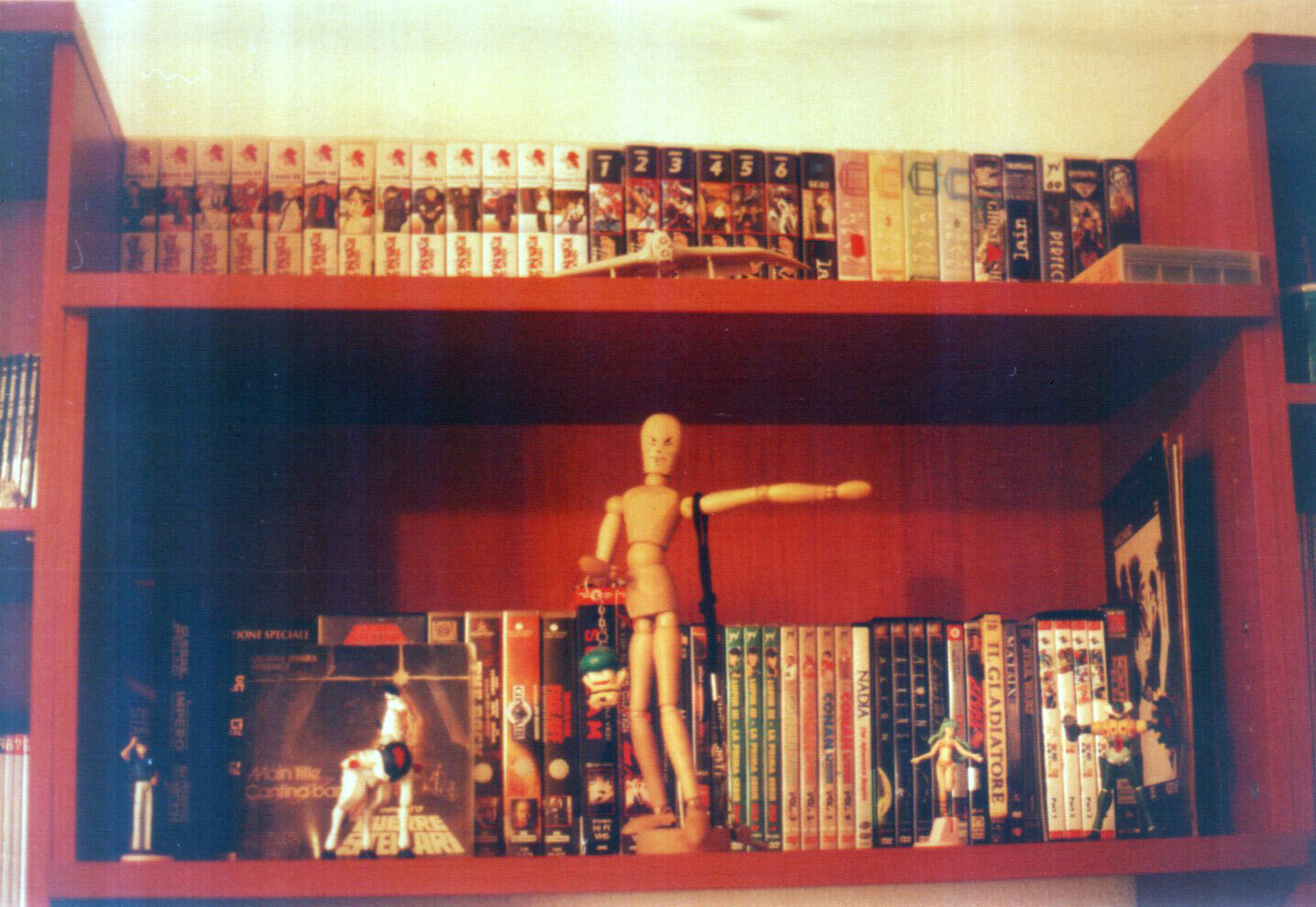 Ripiani di una parete attrezzata, colmi di videocassette e DVD, principalmente di animazione giapponese. Alcune riproduzioni in gomma di personaggi dell'animazione e un manichino di legno al centro. Alcuni dischi in vinile.