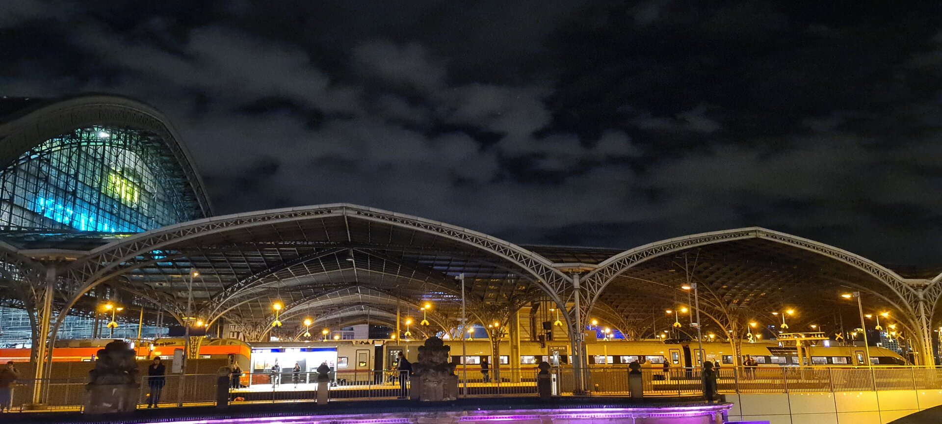 Kölner Hauptbahnhof bei Nacht. Links der von innen beleuchtete Giebel der Haupthalle. Daneben im Rest des Bildes die flachen Bögen der Vorhalle. Viele Lampen beleuchten die Halle mit gelbem Licht. Am dunklen Himmel reflektieren Schleierwolken das Stadtlicht. Kölner Hauptbahnhof bei Nacht. Links der von innen beleuchtete Giebel der Haupthalle. Daneben im Rest des Bildes die flachen Bögen der Vorhalle. Viele Lampen beleuchten die Halle mit gelbem Licht. Am dunklen Himmel reflektieren Schleierwolken das Stadtlicht.