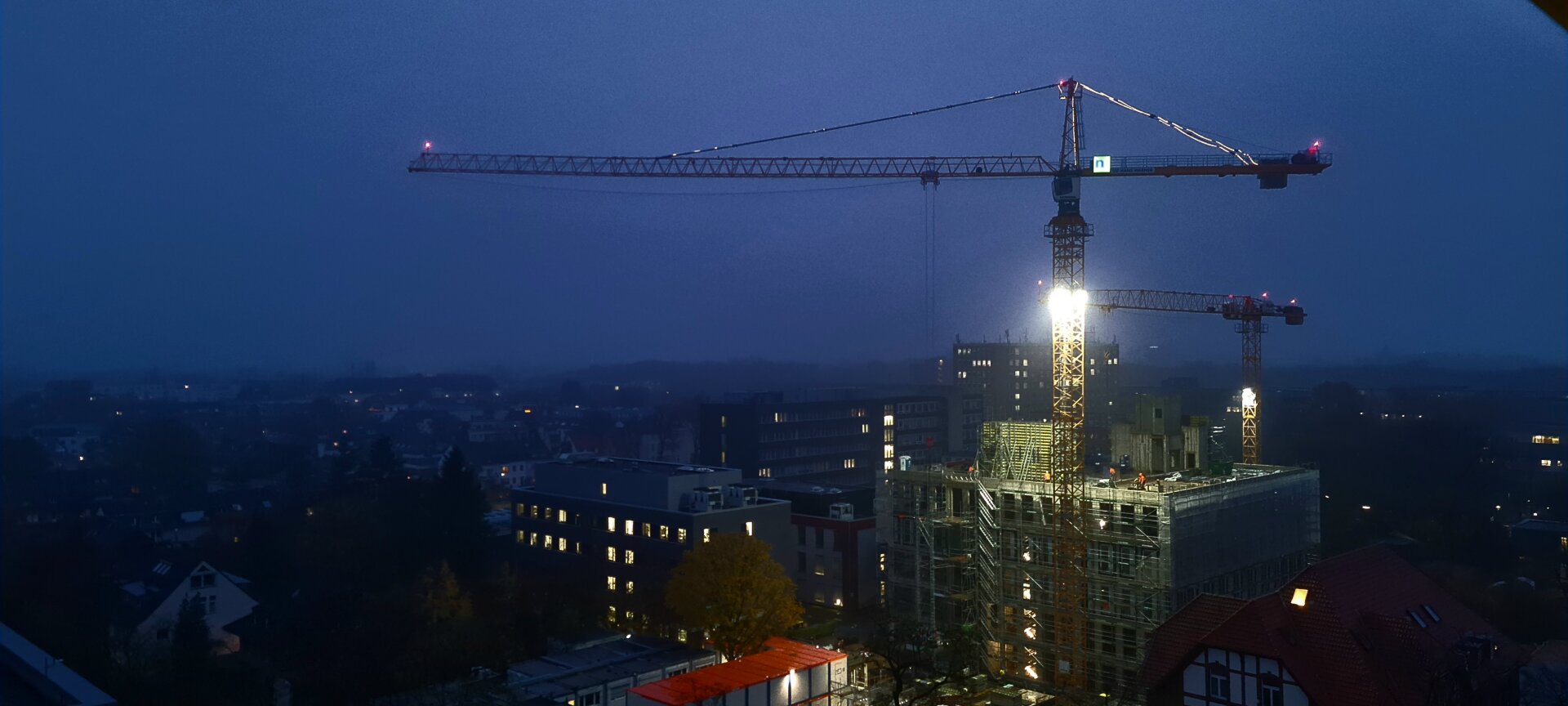 Baustelle eines großen Gebäudes am frühen Morgen. Eine helle Lampe an einem Baukran erhellt die Szenerie.