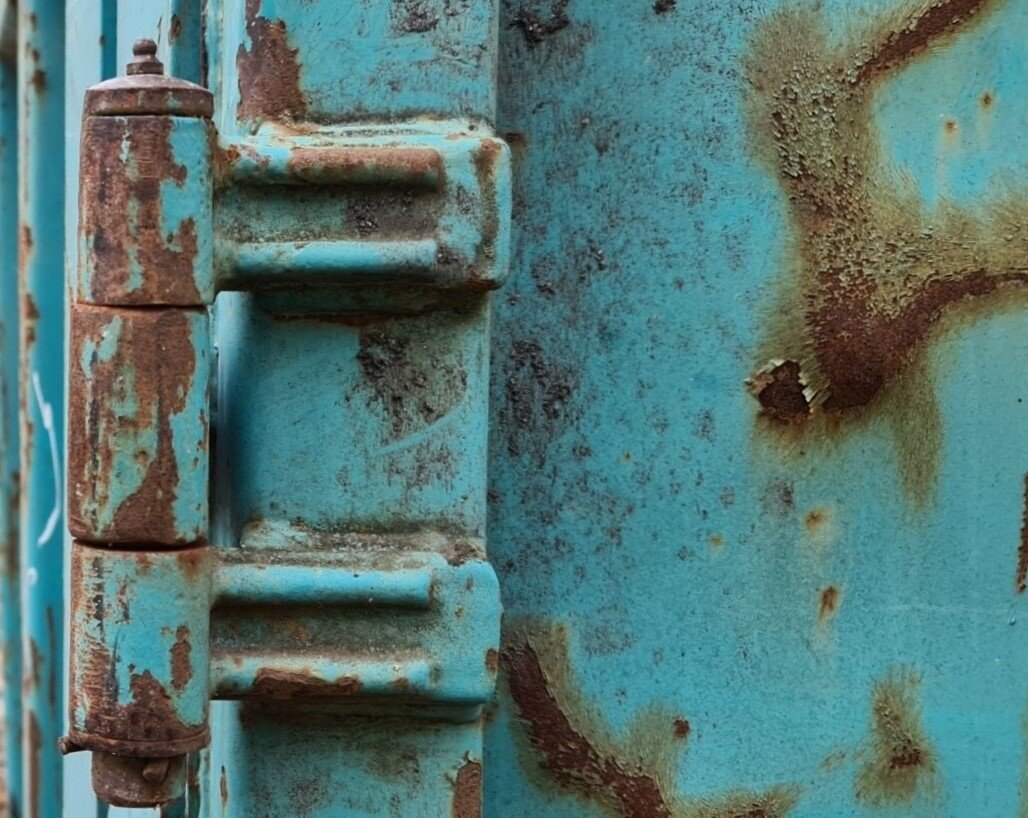 Großes Scharnier an einem blau angestrichenen Container. Die Farbe ist zu großen Teilen abgeblätter und das Eisen verrostet.