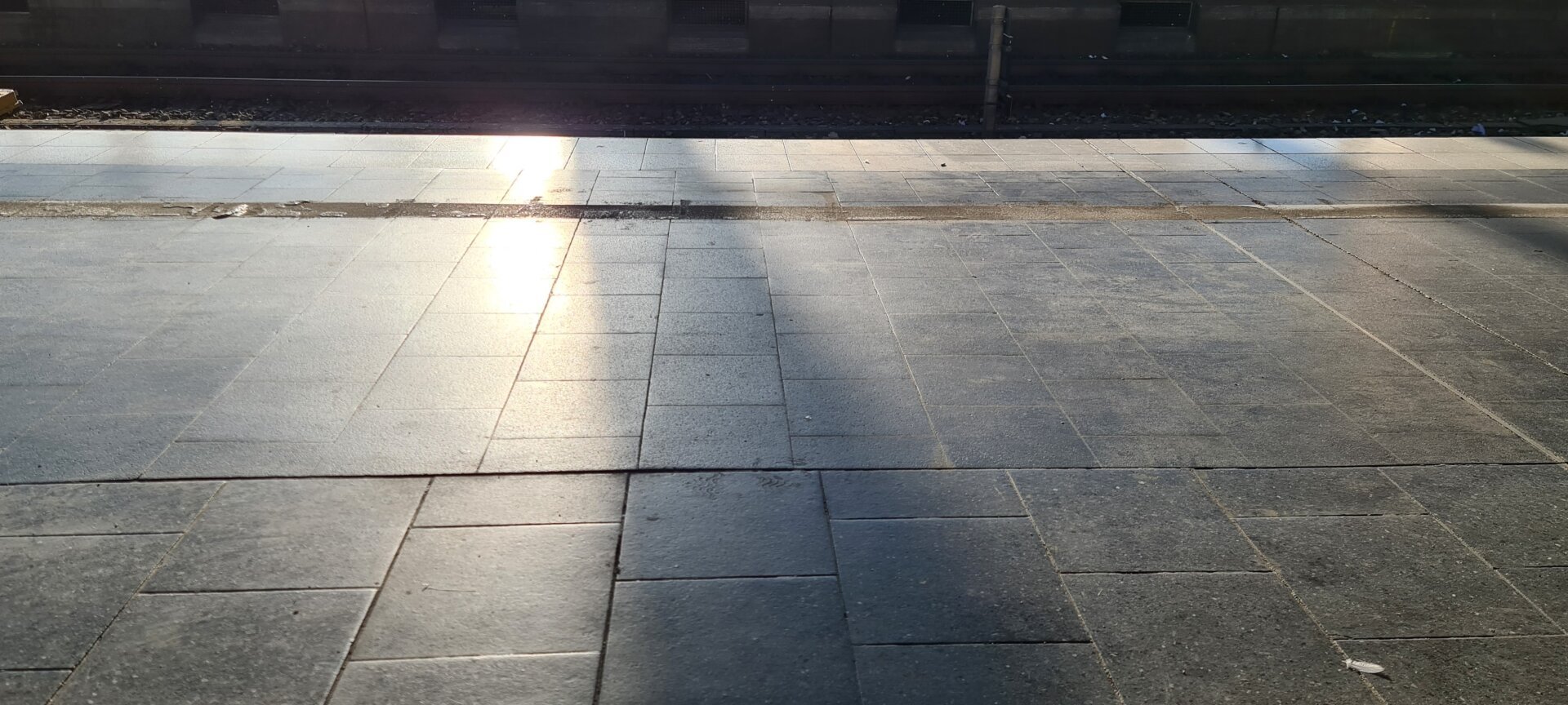 Bodenpflaster eines Bahnsteigs. Im Gegenlicht spiegelt sich das Sonnenlicht. der Schatten eines Pfeilers läuft vertikal durchs Bild. Dahinter liegt im Dunkeln das Gleis und der gegenüberliegende Bahnsteig, sodass das Bild wie eine Schichtung von verschiedenen Graustufen erscheint.