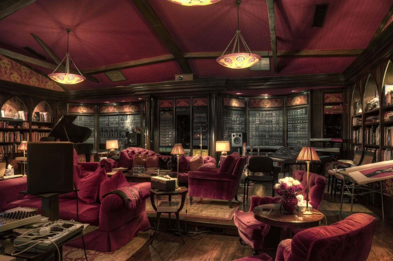 A luxurious music studio with red velvet furniture, bookshelves, and multiple audio equipment. Dim lighting highlights ornate chandeliers and rustic wooden flooring.