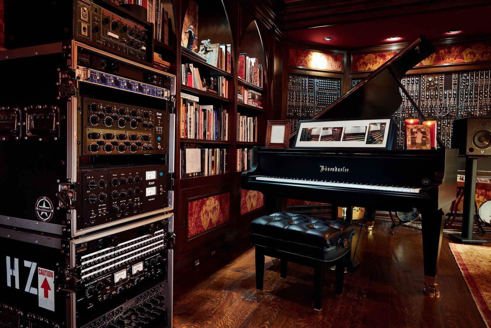 A grand piano in a richly decorated music studio with wooden floors and shelves filled with books and audio equipment.