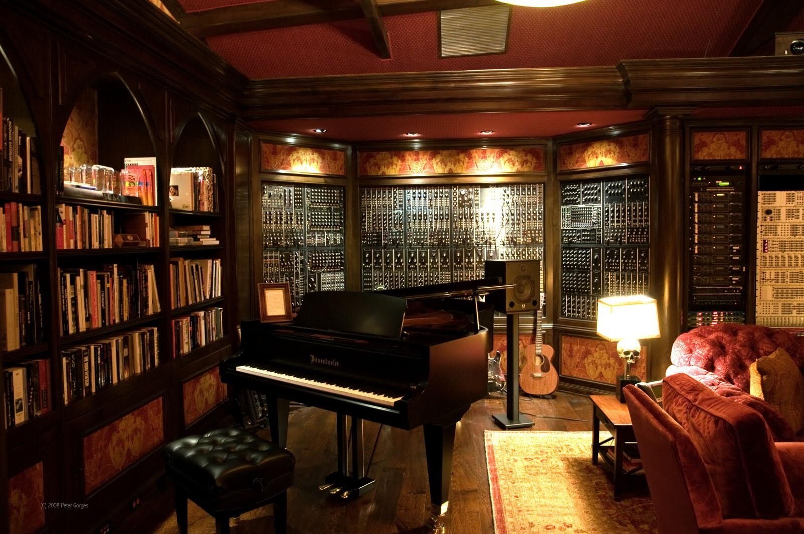 A luxurious music studio with a grand piano, modular synthesizers, bookshelves, a guitar, and comfortable seating with warm lighting and wood accents.