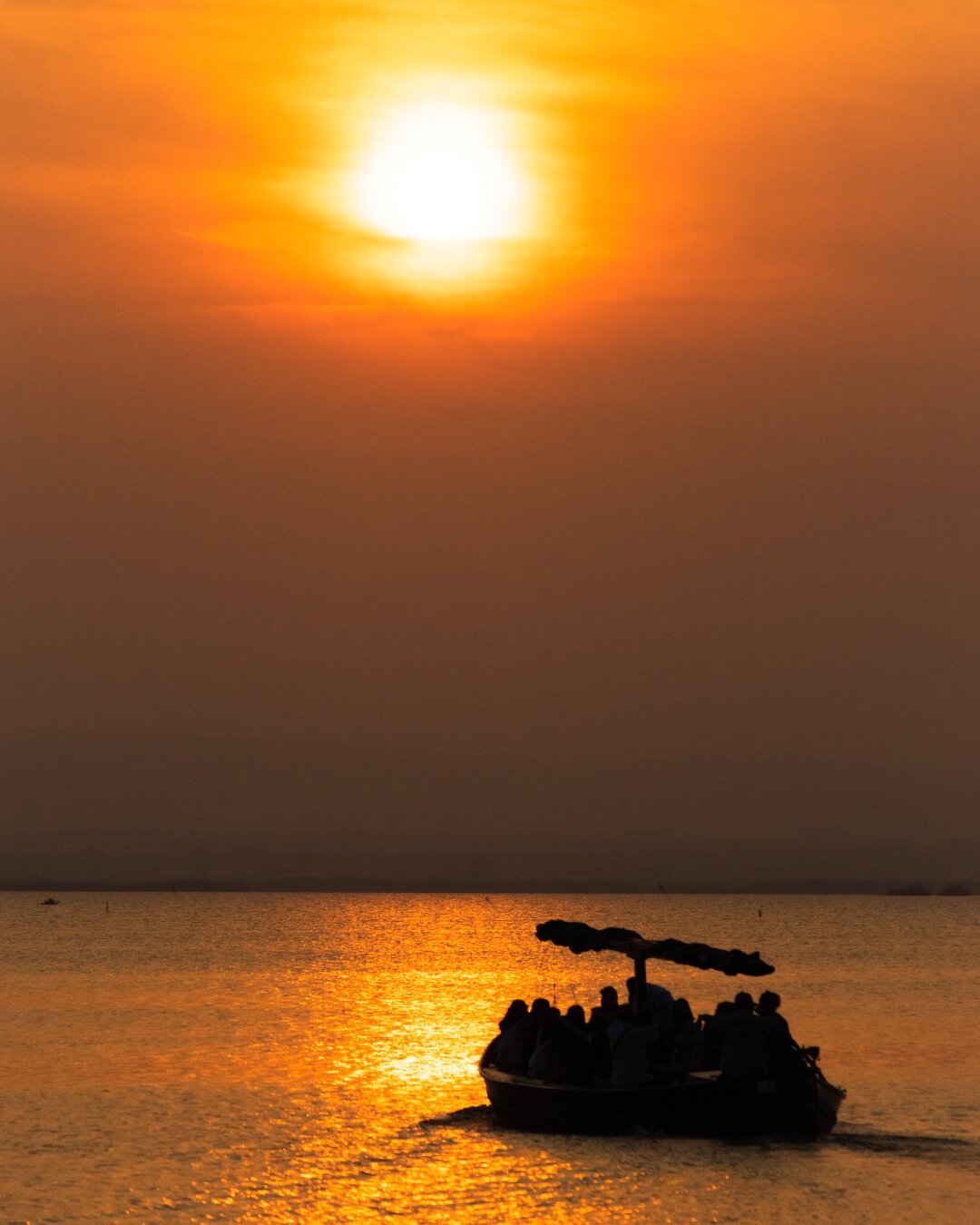 A photo of the sunset with the sun in the background casting an intense orange light over a mass of water and a small ferry sailing to the horizon.