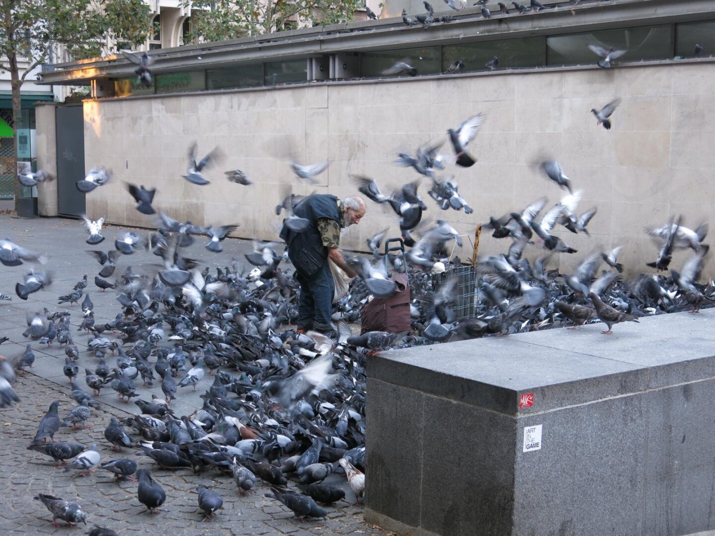 In a small area near the Pompidou, an older man is stooped over and reaching into a bag of bird seed. He is completely surrounded by hundreds of pigeons, both on the ground, and flying around him.