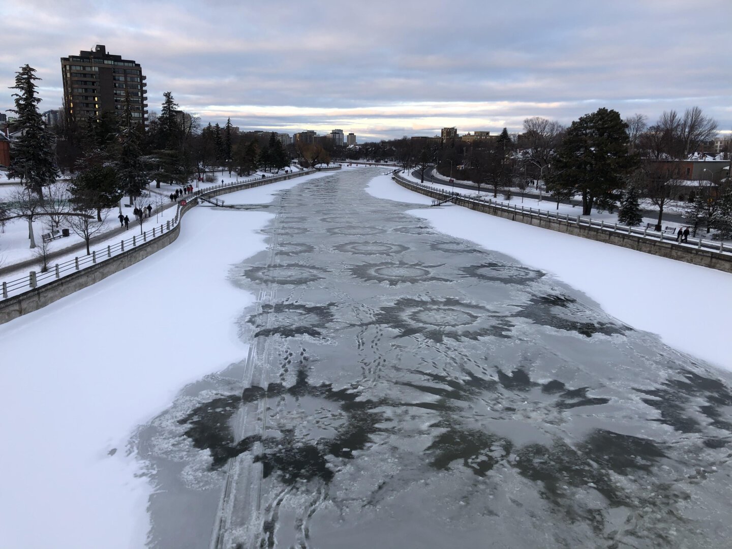 I'm standing on a pedestrian bridge looking down the nearly frozen canal. There are many unusual star-shaped patterns in the ice that have formed, I've heard, because the canal maintenance crew has drilled holes in the ice to allow for flooding. The background is mainly trees along with apartment and office buildings.