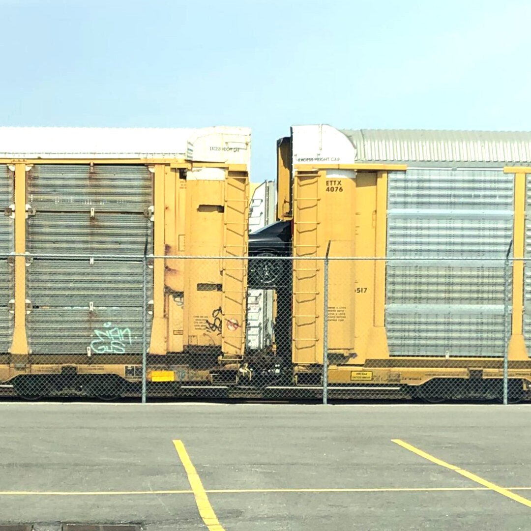 From across an empty parking lot, two railway car carriers. Newly built cars are being loaded onto the train and there is a black Dodge Challenger that is currently in between the two railway carriers as its moving along to where it will be parked for shipping.