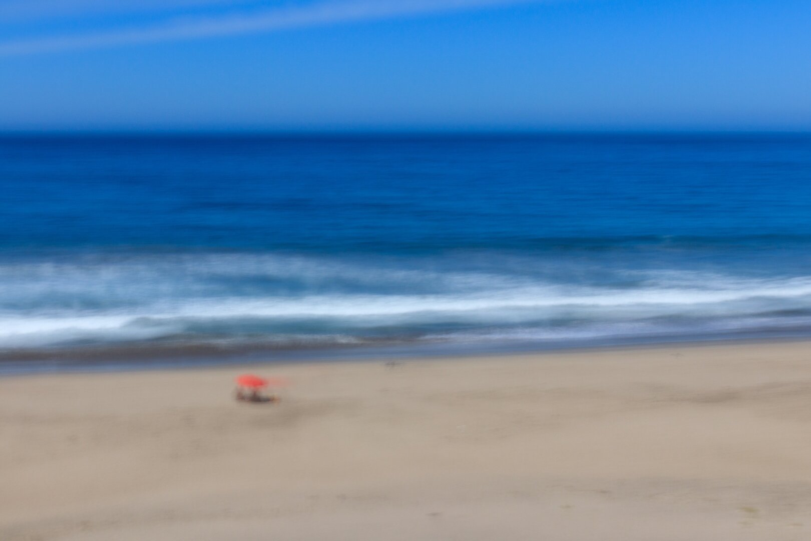 Español: fotografía desenfocada de una playa vacía con un sombrilla roja pequeña junto a la orilla, cielo azul despejado y olas rompiendo suavemente en la arena.

English: blurred photo of an empty beach with a small red umbrella near the shoreline, clear blue sky, and waves gently breaking on the sand.