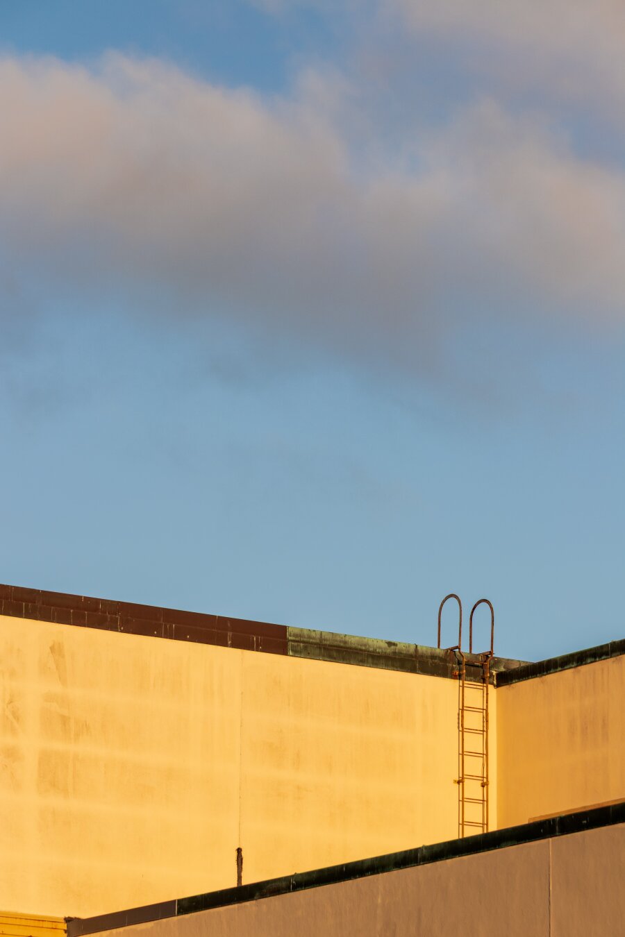 Español: fachada de un edificio amarillo iluminado por la luz dorada del atardecer, con una escalera metálica en el tejado bajo un cielo azul con nubes suaves.

English: yellow building exterior bathed in golden sunset light, featuring a roof ladder against a clear blue sky with soft clouds.