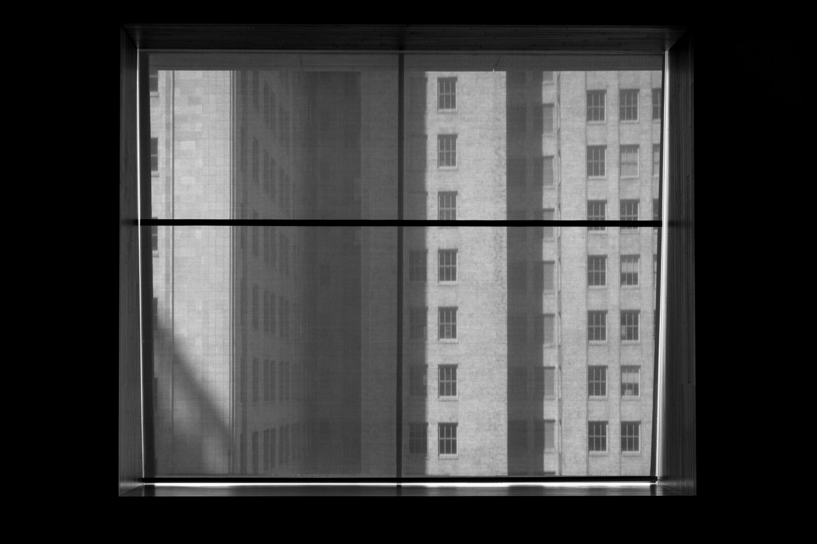 Español: fotografía en blanco y negro a través de una ventana. Se ven las fachadas de edificios urbanos con ventanas rectangulares tras una persiana traslúcida que suaviza la escena.

English: black and white photo looking through a window. High-rise building facades with rectangular windows are visible through a semi-transparent blind.