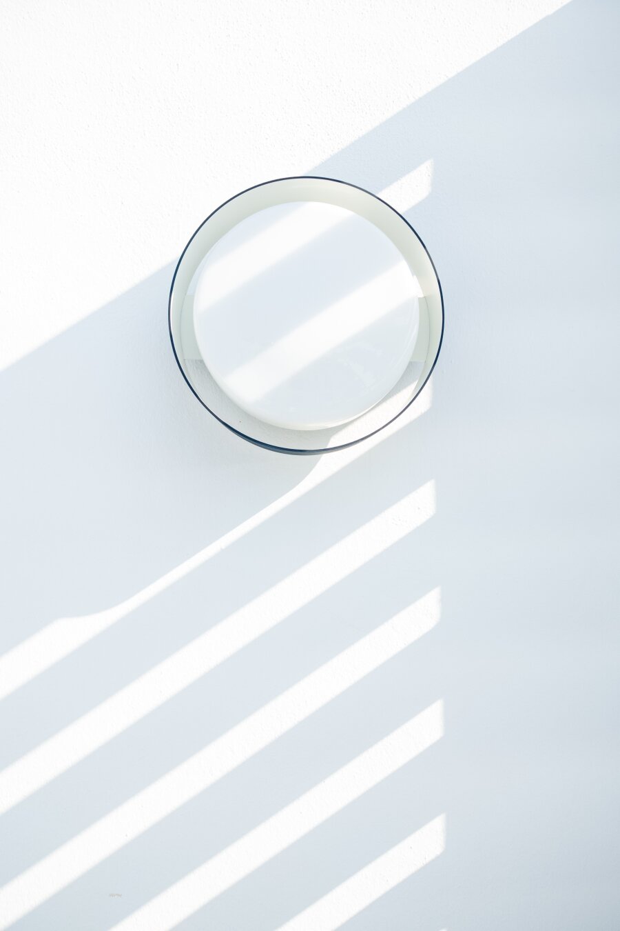 Español: vista superior de un plato blanco redondo con borde oscuro sobre una superficie blanca brillante. La luz del sol ilumina la escena en diagonal, creando sombras paralelas en forma de persiana que atraviesan la superficie y el plato. Estilo minimalista y limpio.

English: top-down view of a round white plate with a dark rim resting on a bright white surface. Sunlight streams diagonally, casting parallel slatted blind shadows across the surface and the plate. Minimalist and clean aesthetic.
