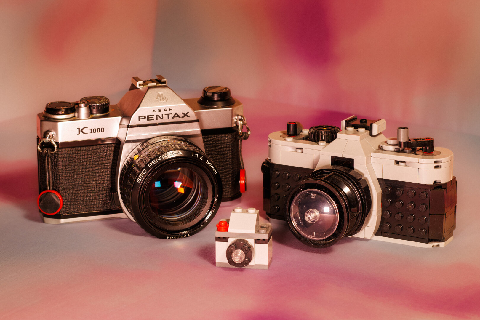A family portrait of three cameras. A Pentax K1000, a smaller LEGO camera, and a very small LEGO camera. On a swirly pastel pink background. Like a cheesy family portrait.