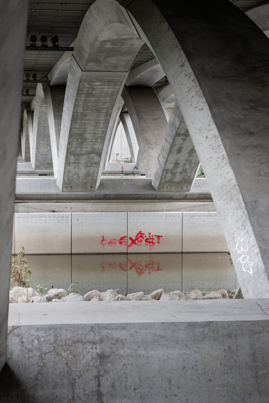 "Coexist" in different symbols, in red, on the bottom pylon of a bridge over a river, reflected in the water.
