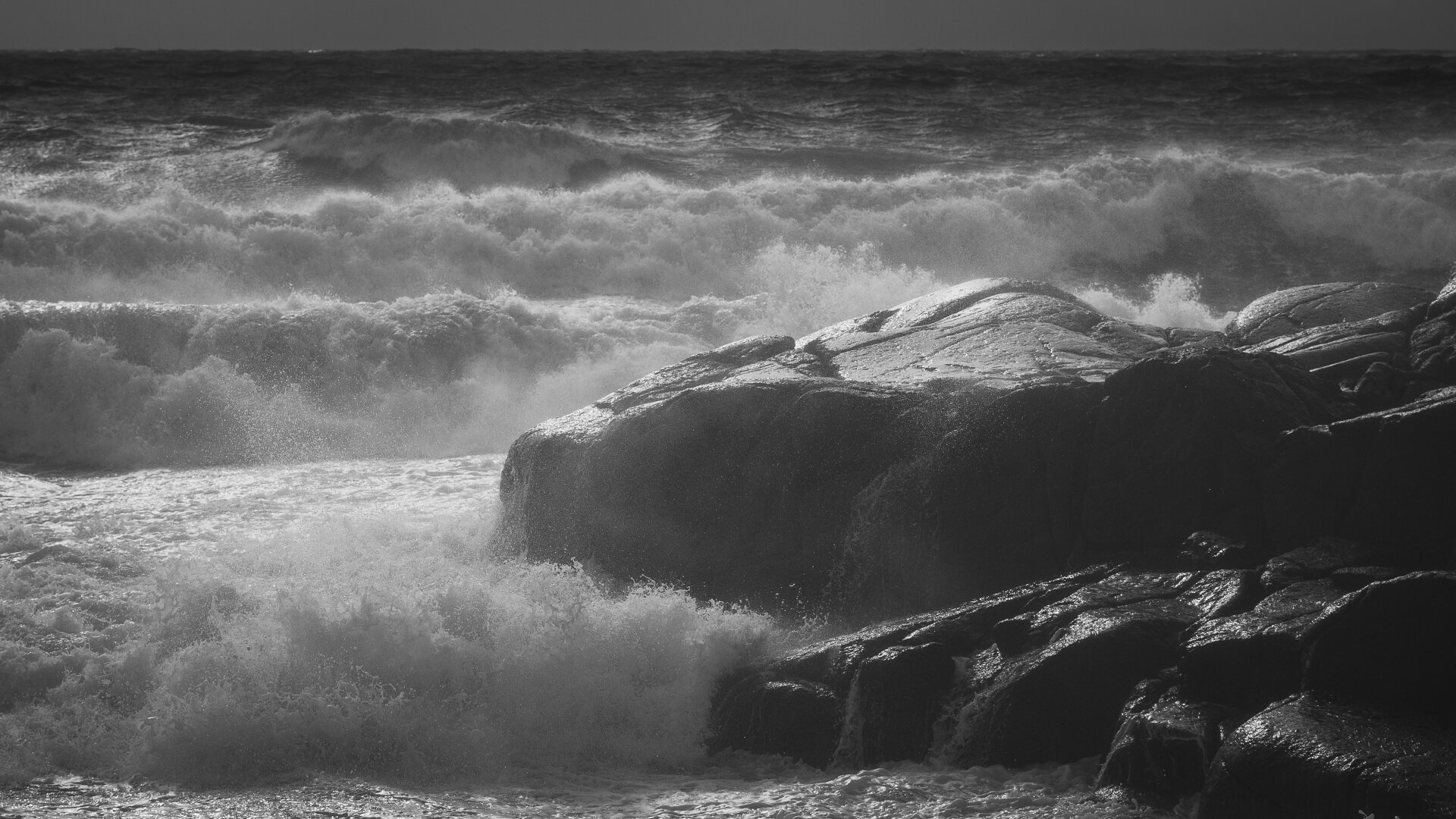 A wide aspect ratio monochrome photograph of large waves on a stormy day around a black rock.

The wave dominates the left side of the image, sunlit and white, forming a mass of foamy breaking waves, backlit by sunlight. The rock dominates the right side of the image, dark and wet, glistening on the weather worn top side from the sunlight.

In the background is a view of the sea, darker toward the horizon. A small strip of sky is visible at the very top of the photograph.