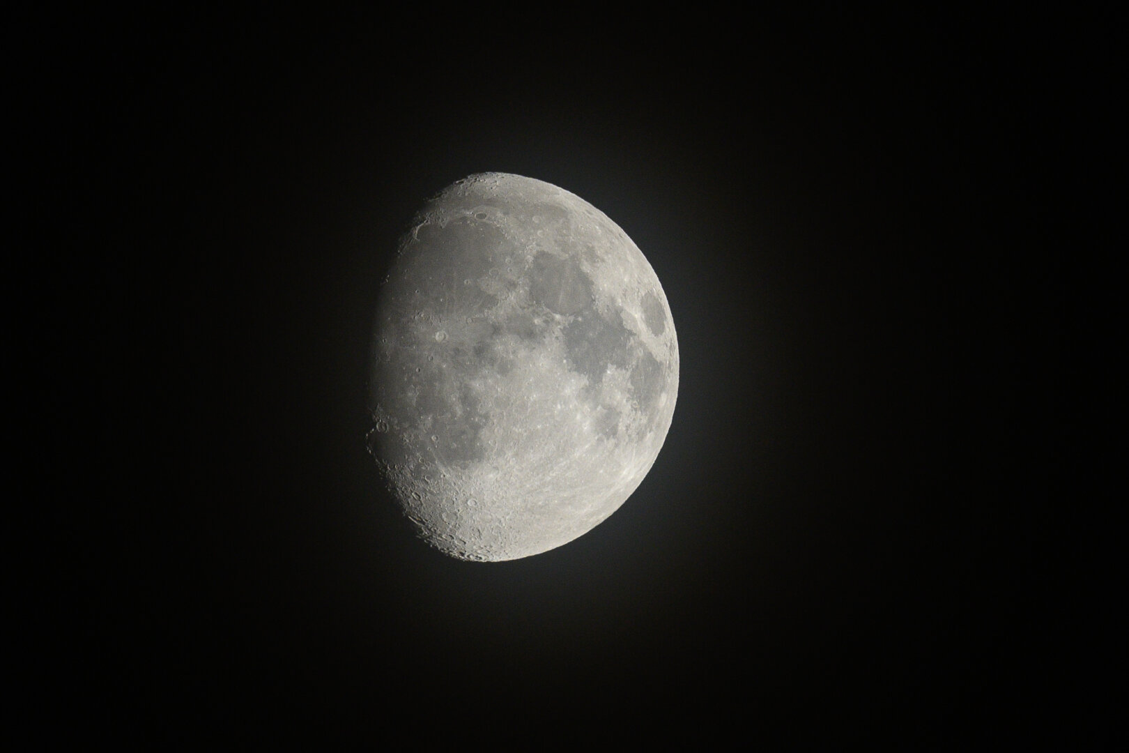 A photograph of the moon, about two-thirds full. The moon is slightly to the left of center and fills a quite large portion of the frame, the rest of which is pitch black, except for a lisght haze around the moon from a var light cloud cover.